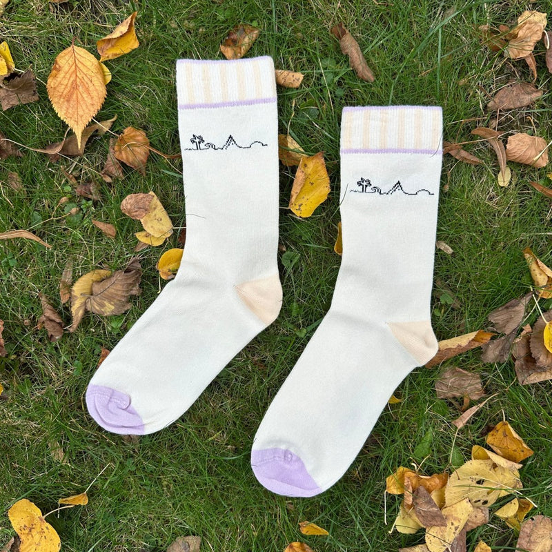 Insel-Bio-Baumwollsocken: Der Sockensaum ist ca. 4cm breit und mit vertikal laufenden Streifen in beige versehen. oberhalb und unterhalb  angrenzend an die Vertikal-Streifen verläuft ein Querstreifen in lila. Das Druck-Motiv der Socke liegt zwischen dem Saum und der Ferse und heißt "Insel": es besteht von links nach rechts aus: Palme, Welle, Berg und aufgehender Sonne. Die einzel. Symbole sind durch e. Linie durchgehend verbunden. Der Druck ist schwarz. Ferse ist beige; Sockenspitze ist lila. Foto Ungerade