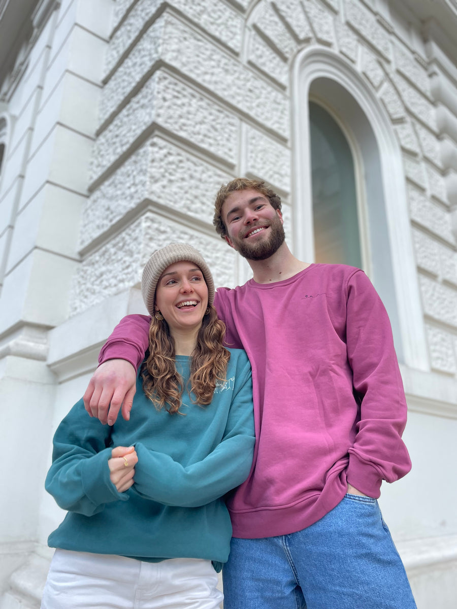 Frau (links) und Mann (rechts) tragen Unisex Oversized Sweatshirt. Frau trägt grün mit Catband Motiv, mann trägt rot/rose mit Insel-Motiv. Beide Motive sitzen über der linken Brust. Der Motiv-Druck ist beim grünen Sweatshirt weiß, beim rose farbenen Sweatshirt schwarz. Foto von Ungerade Wien