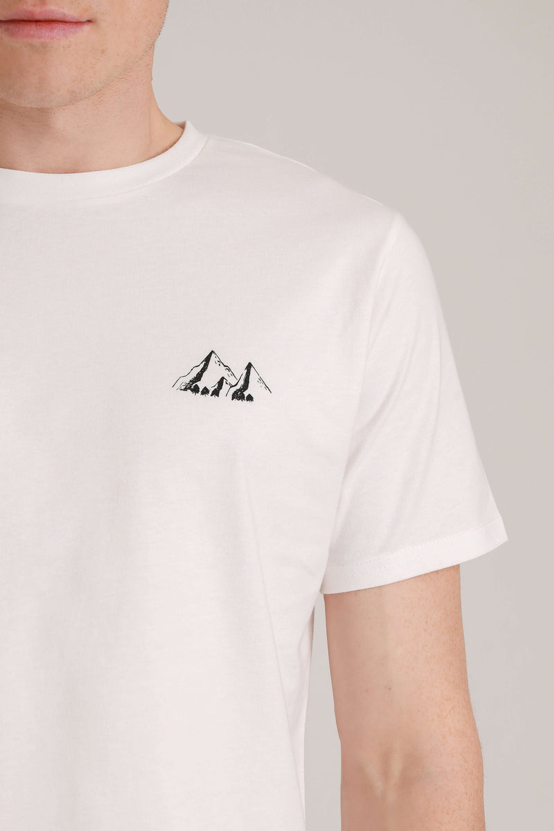 Close Up Foto von weißem Shirt  mit Berge-Motiv (schwarzer Druck). Das Berge-Motiv befindet sich über der linken Brust des Shirt-Trägers. Das Motiv besteht aus 3 Bergen: 
von links nach rechts: beginnend mit einem großen Berg, daneben versetzt nach vorne ein kleiner Berg und ein großer Berg rechts daneben. Die Berge sind alle verbunden. Das kleine Extra sind die winzigen Bäume (Tannen), die sich am Fuß der Berge befinden. Die Berge sind schattiert, sodass Tiefe und Kontur entsteht. 