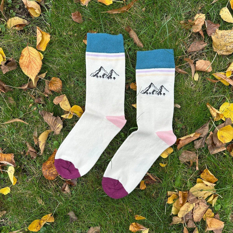Mountain Peaks – Bio-Baumwollsocken für Gipfelstürmer & Freiheitsliebende: Der dunkelgrüne Saum ist 4cm breit. Darunter befinden sich zwei Querstreifen in Ilia und beige. ca. 2cm darunter befindet sich das Berg-Motiv (3 Berge - groß, klein, groß - schattiert, am Bergfuß sind Tannen gezeichnet. Der Druck ist schwarz. die Sockenferse ist hellrosa, die Sockenspitze dunkelrot. Die Socken liegen im grünen Gras, umgeben von Herbstblättern. Foto von Ungerade Wien
