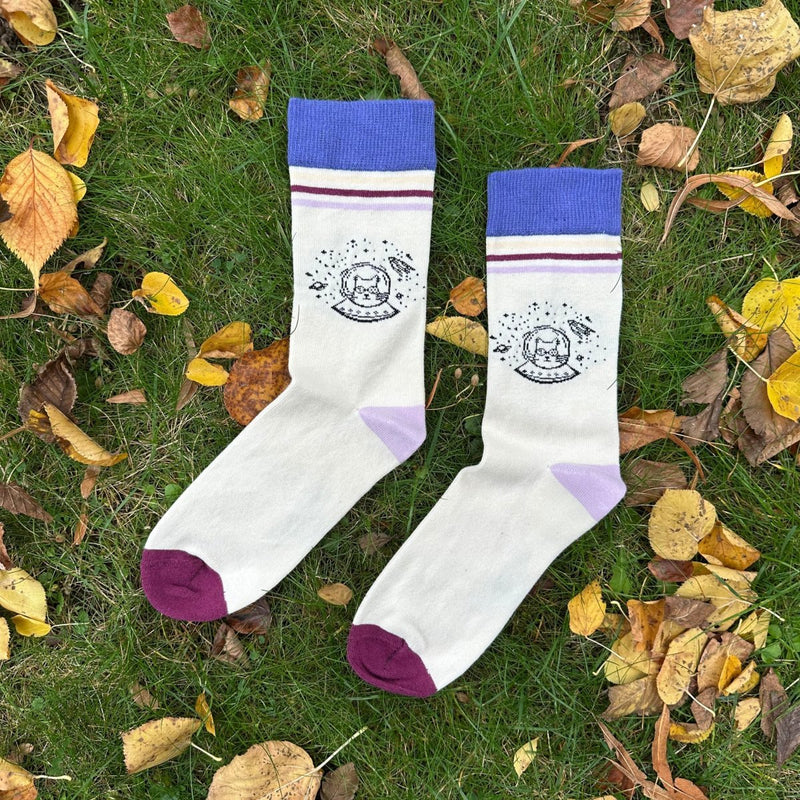 Space Cat Stripe – Bio-Baumwollsocken mit galaktischem Katzen-Design. Die Socken liegen im grünen Gras, umgeben von Herbstblättern. Der Sockenbund ist ca. 4cm breit. Darunter liegen 3 Querstreifen (3-5mm dick) in hellgelb, dunkelrot und lila. darunter das Space-Cat Motiv: Ein Katzenkopf mit Spacebrille in einem Space-Helm und halbem Ufo. rechts, links und über dem Kopf befinden sich Sterne (Punkte), eine kleine Rakete und Planeten. Die Ferse der Socke ist lila, die Sockenspitze dunkelrot. Ungerade Wien