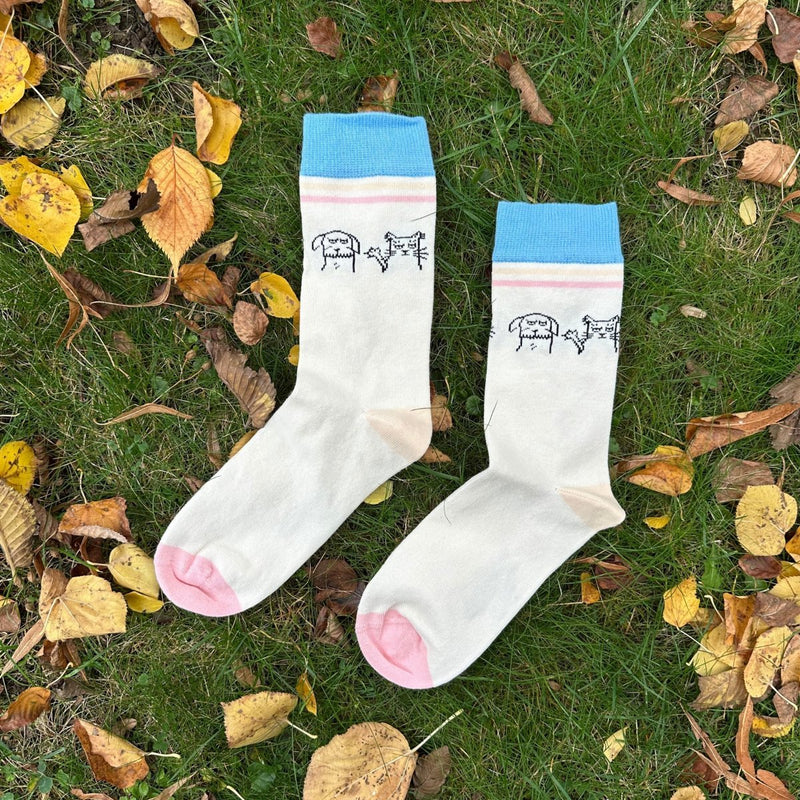 Cat & Dog Friends – Bio-Baumwollsocken über Freundschaft mit Miau und Wuff: Der Saum ist ca. 4cm breit und hellblau. Unter dem Saum liegen 2 Querstreifen in beige und rosa. Darunter liegt das gezeichnete Druckdesign: Ein Hund links (schaut grimmig), die grantige Katze rechts daneben mit hochgehaltener Pfote und ausgefahrenen Krallen. Die Ferse ist hellbeige, die Sockenspitze ist rosa. Das Sockenpaar liegt auf grünem Gras, umgeben von Herbstblättern  - Ungerade Wien