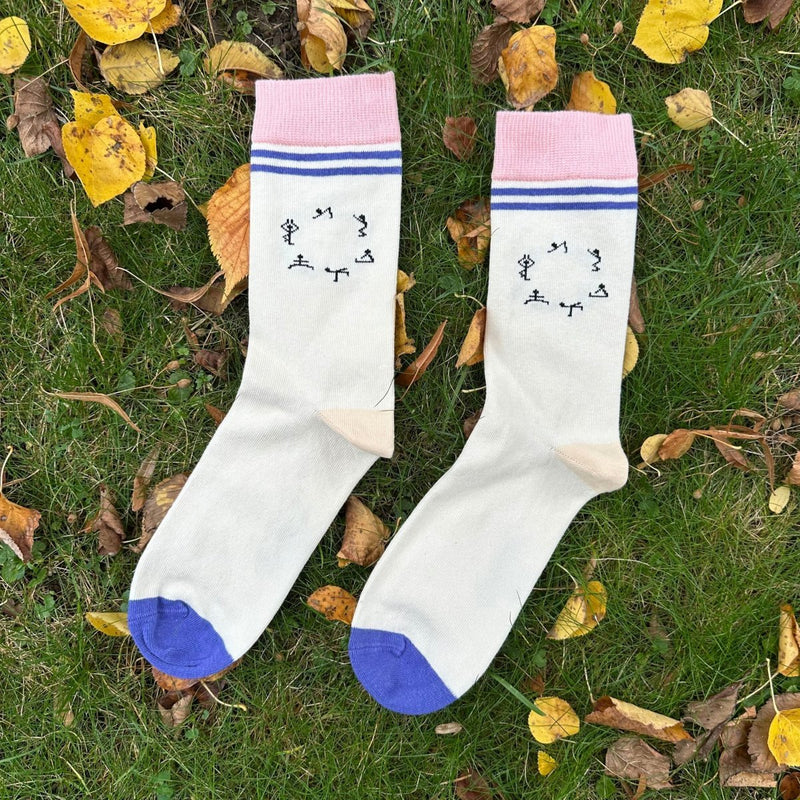 Yoga – Bio-Baumwollsocken für Balance, Bewegung & innere Stärke. 
Der Sockensaum ist ca. 4cm breit und hellrosa. Unter dem Saum befinden sich zwei Querstreifen, beide dunkellila. 2cm darunter befindet sich das Yoga-Motiv: 6  Strichmännchen im Kreis angeordnet mit unterschiedl. Yogaposen.  Die Ferse ist hellbeige, die Sockenspitze dunkellila. Das Sockenpaar liegt auf grünem Gras, umgeben von Herbstblättern. Foto von Ungerade Wien